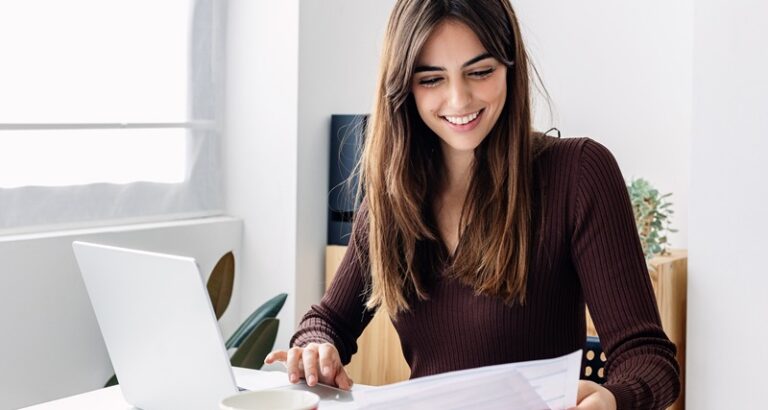 Young smiling pretty adult woman working on laptop while checking home invoices