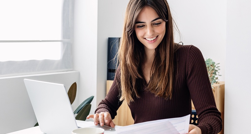 Young smiling pretty adult woman working on laptop while checking home invoices
